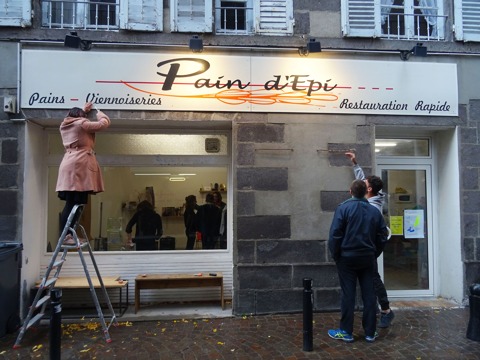On voit la devanture d'n batiement avec l'enseigne Pain d'Epi. Une personne est sur un escabot et enlèvre uen vis de l'enseigne, deux autres personnes regardent d'en bas.