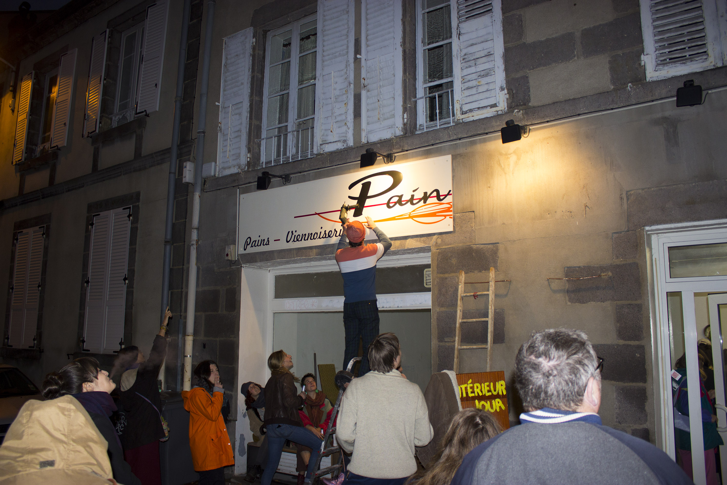 Photo de nuit, un homme est sur un escabot et tente de dévisser la deuxème partie de l'enseigne. Une foulle l'entoure et le regarde depuis la rue. Un bras levé lui indique quelqu'un chose.