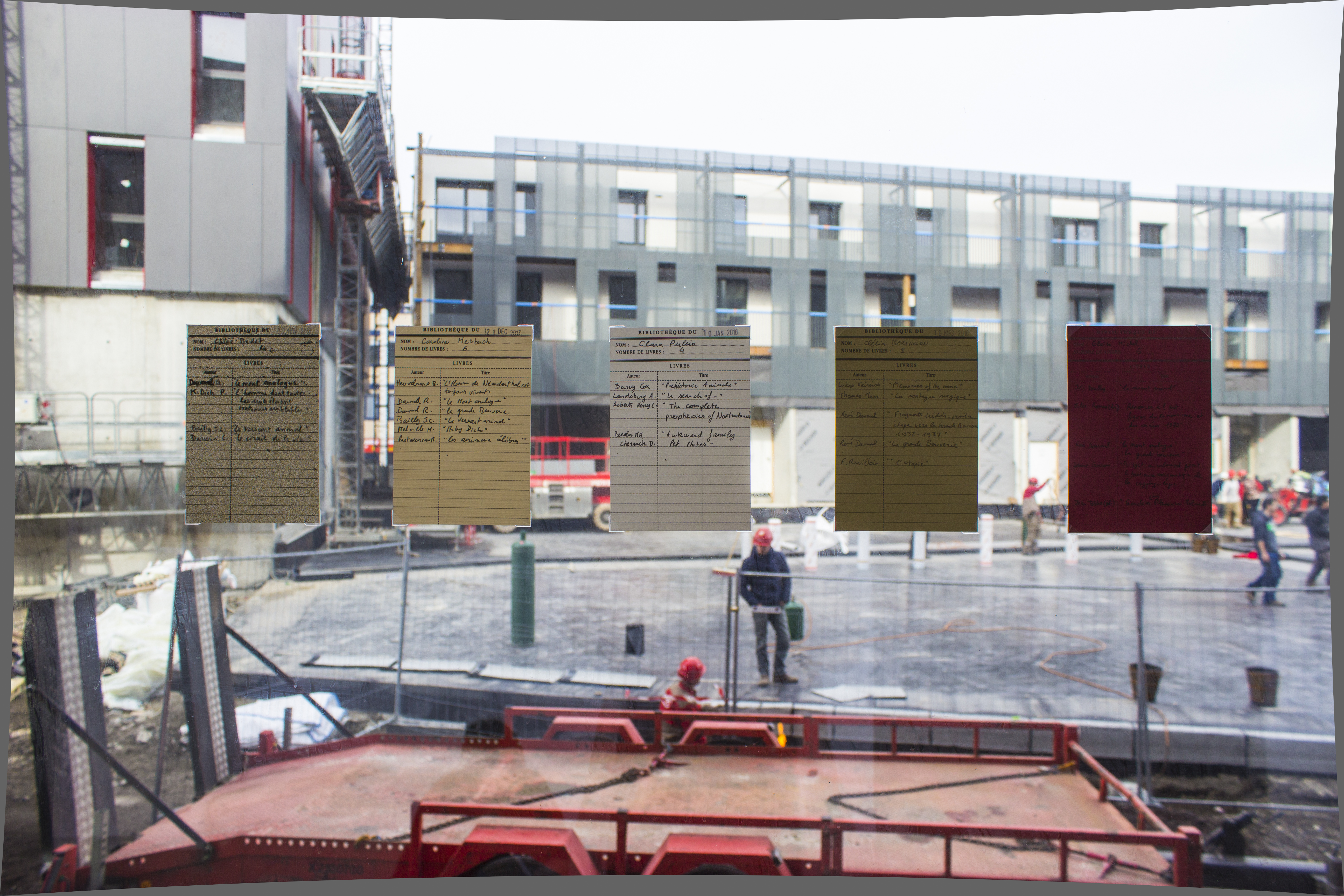 Cinq fiches de bibliothèques sont posées sur une vitre. Derrière on voit un chantier.