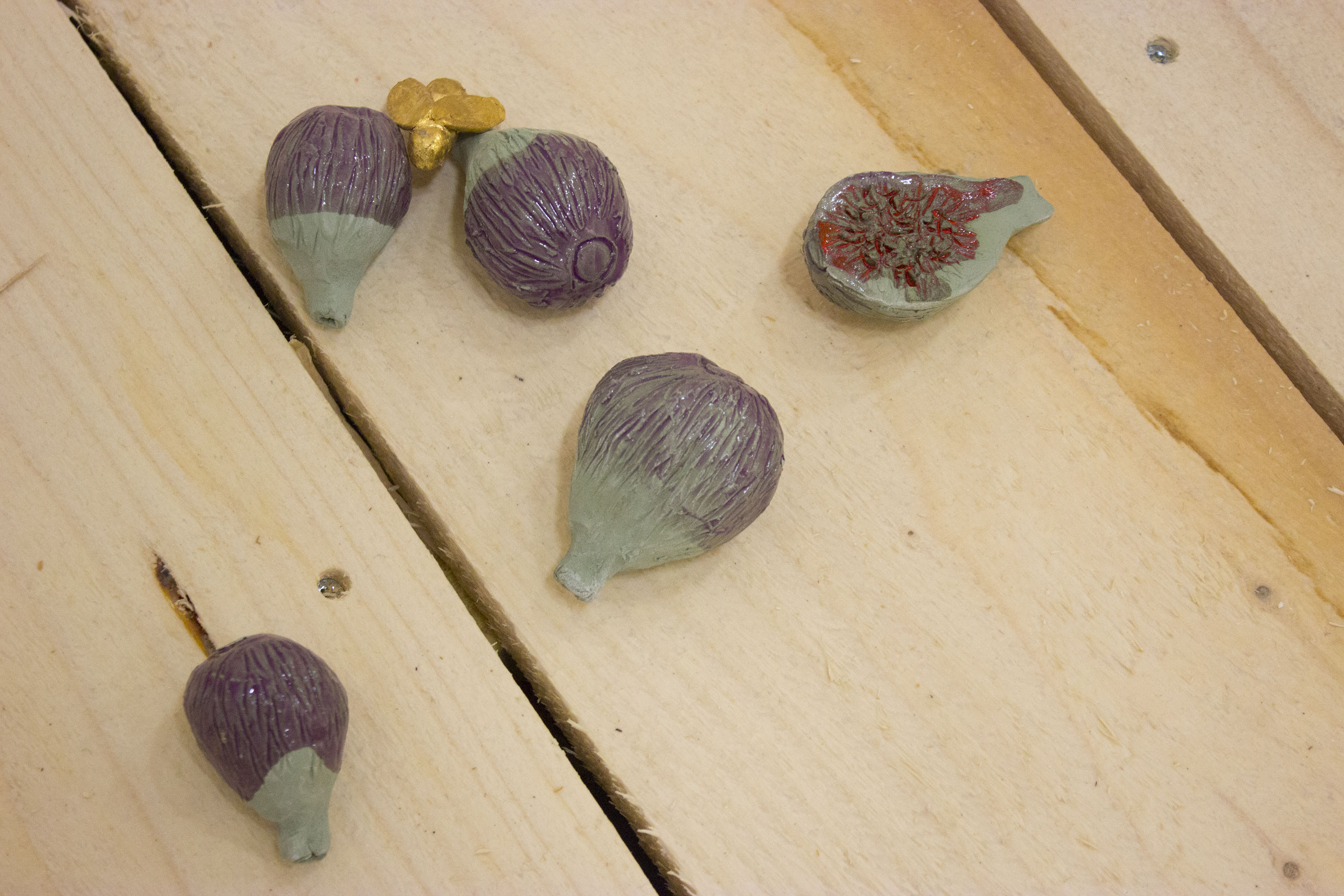 gros plan sur les figues en céramique verte, émaillé en violet transparent posées à même le sol. L'une est ouverte et contient de l'émail rouge. Une guèpe dorée est posée sur une figue.