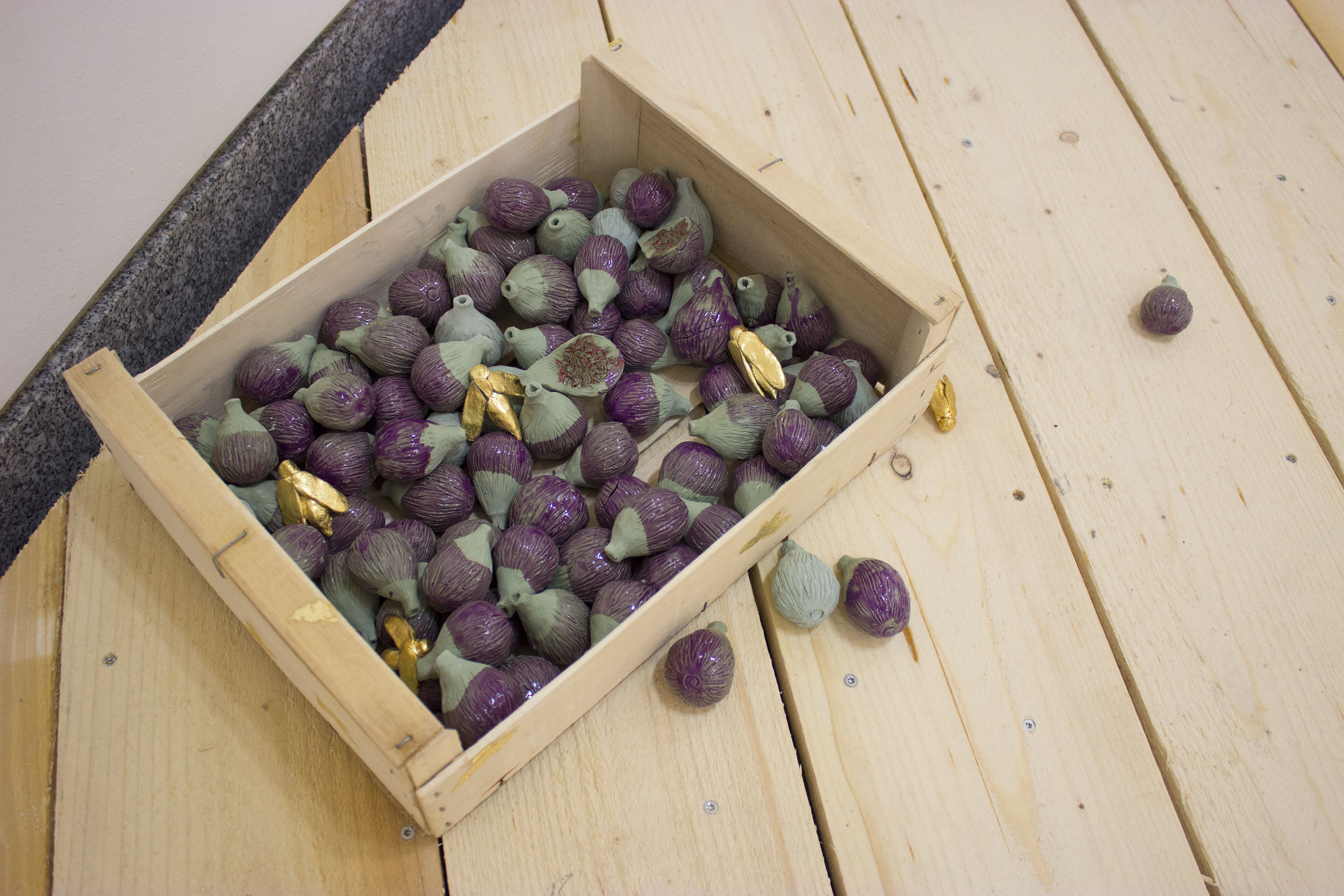 une cagette en bois est posée à même le sol. A l'intérieur, des centaines de figues en céramique verte, émaillées d'un violet transparent. Elles ont toutes des tailles différentes, certaines sont pleines d'autres sont ouvertes en deux. Au milieu, quelques guèpes dorées se sont posées là. Quelques figues sont à l'extérieur de la cagette.