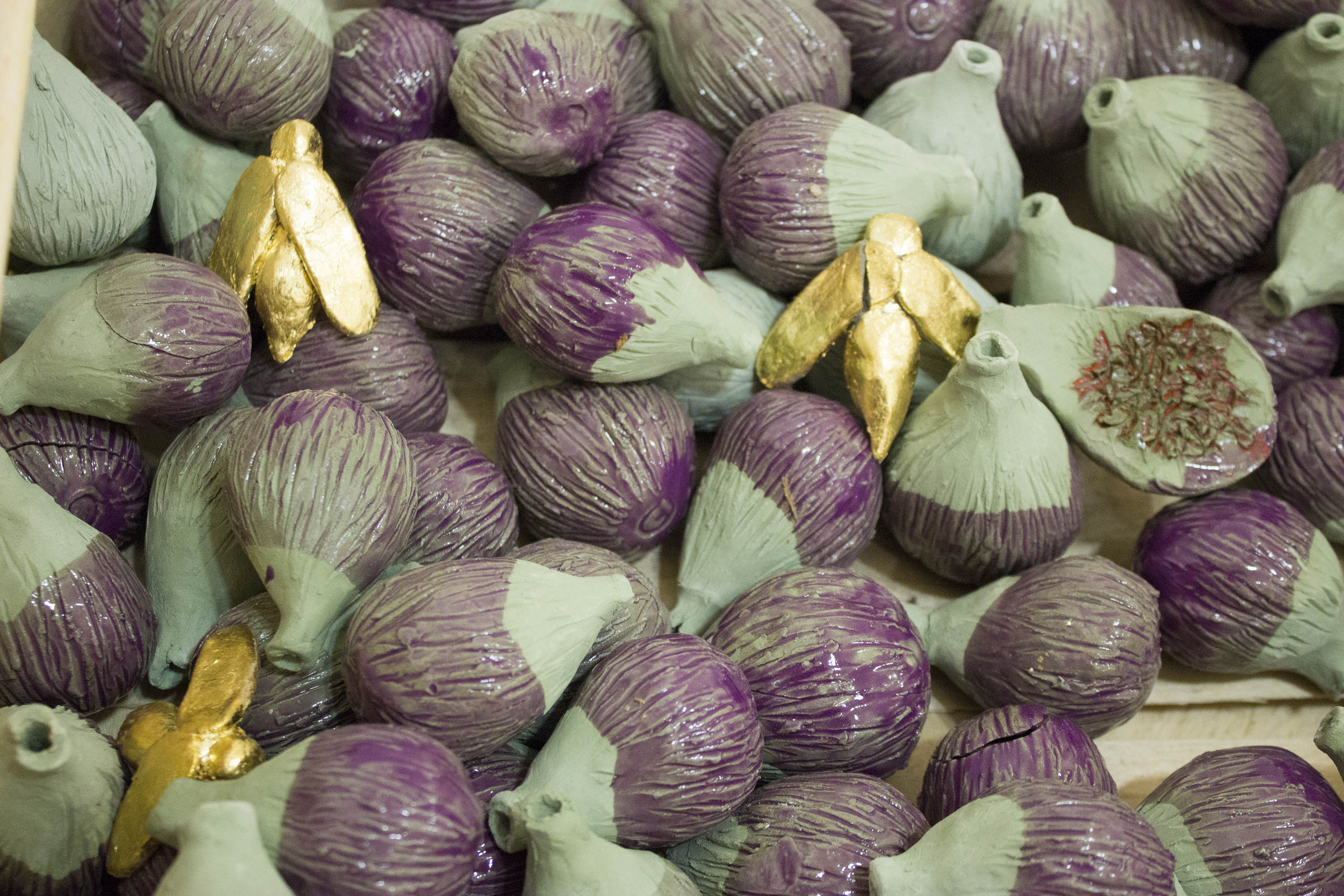 gros plan sur les figues en céramique verte, émaillé en violet transparent dans la cagette. L'une est ouverte et contient de l'émail rouge, d'autres sont presque éclatées. Trois guèpes dorées est posée sur dans la cagette, l'une a une aile cassée.