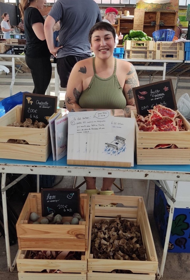 on voit Clélia assise derrière un petit stand dans un marché. Elle est tout sourire.