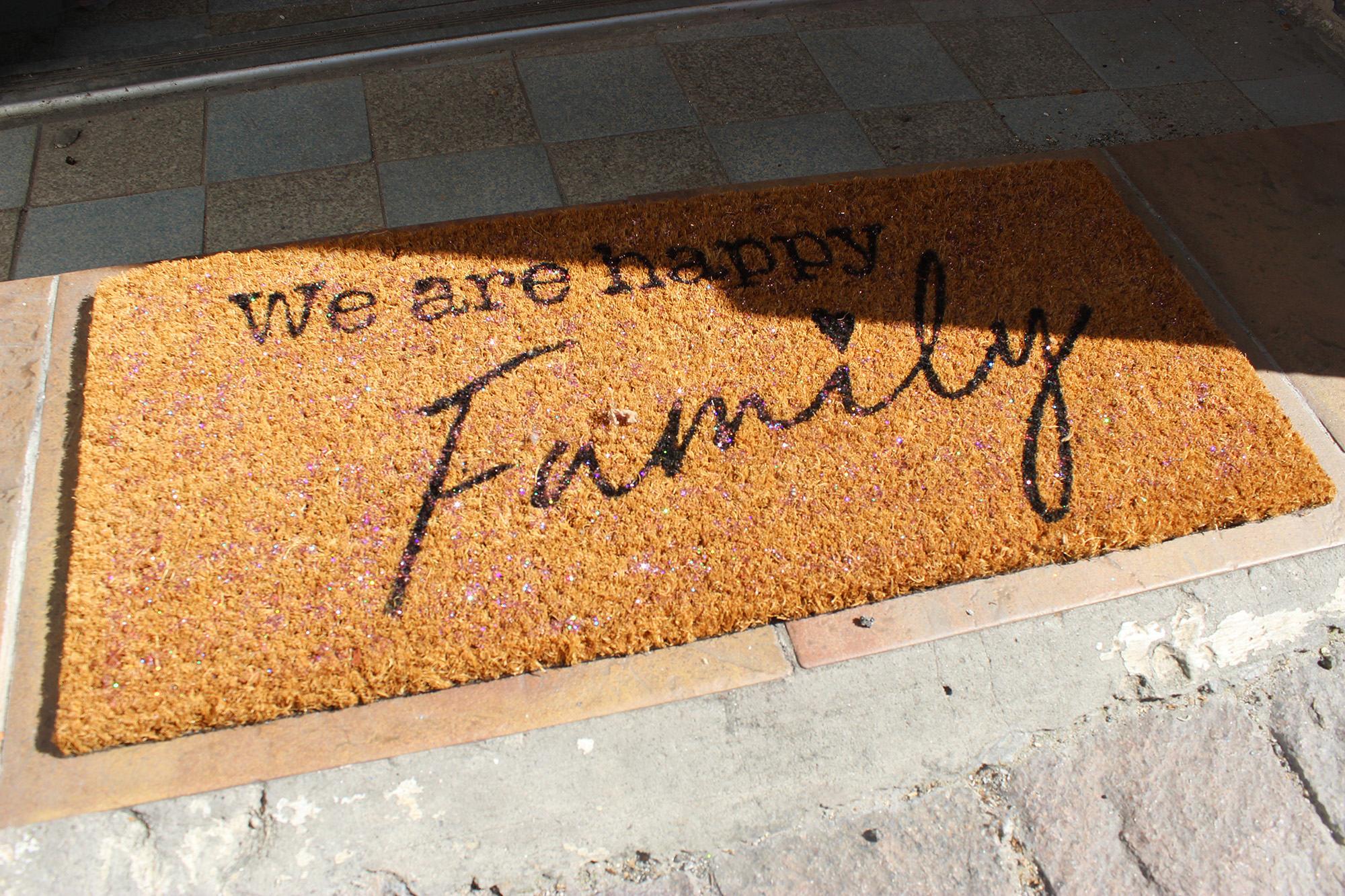 Un paillasson avec l'inscription We are happy Family est posé à l'entrée d'une porte. On apperçoit des paillettes à l'intérieur.