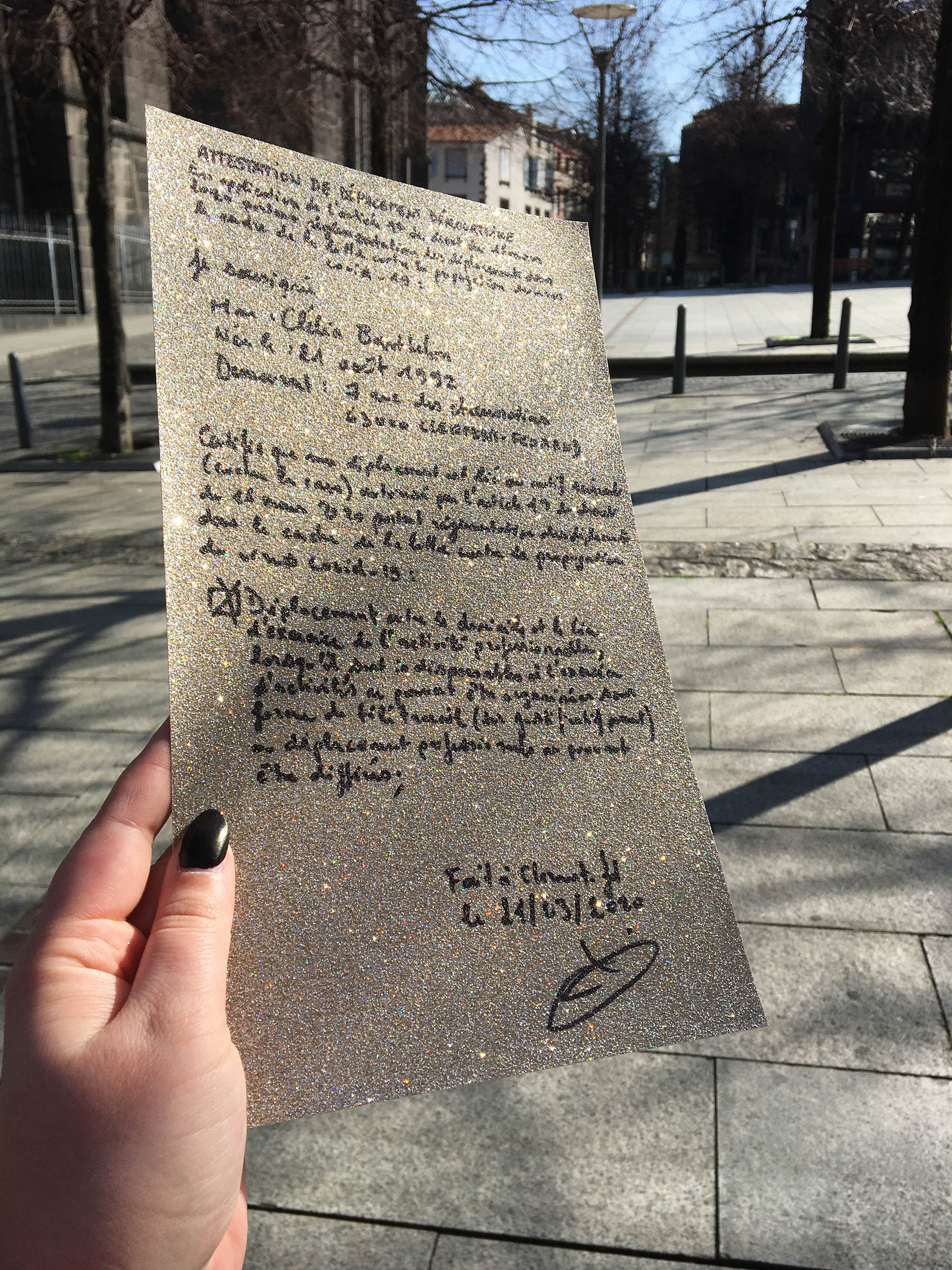 Photo prise à l'extérieur, sur une feuille pailletée on voit écrit à la main une attestation de déplacement dans la periode du Covid.