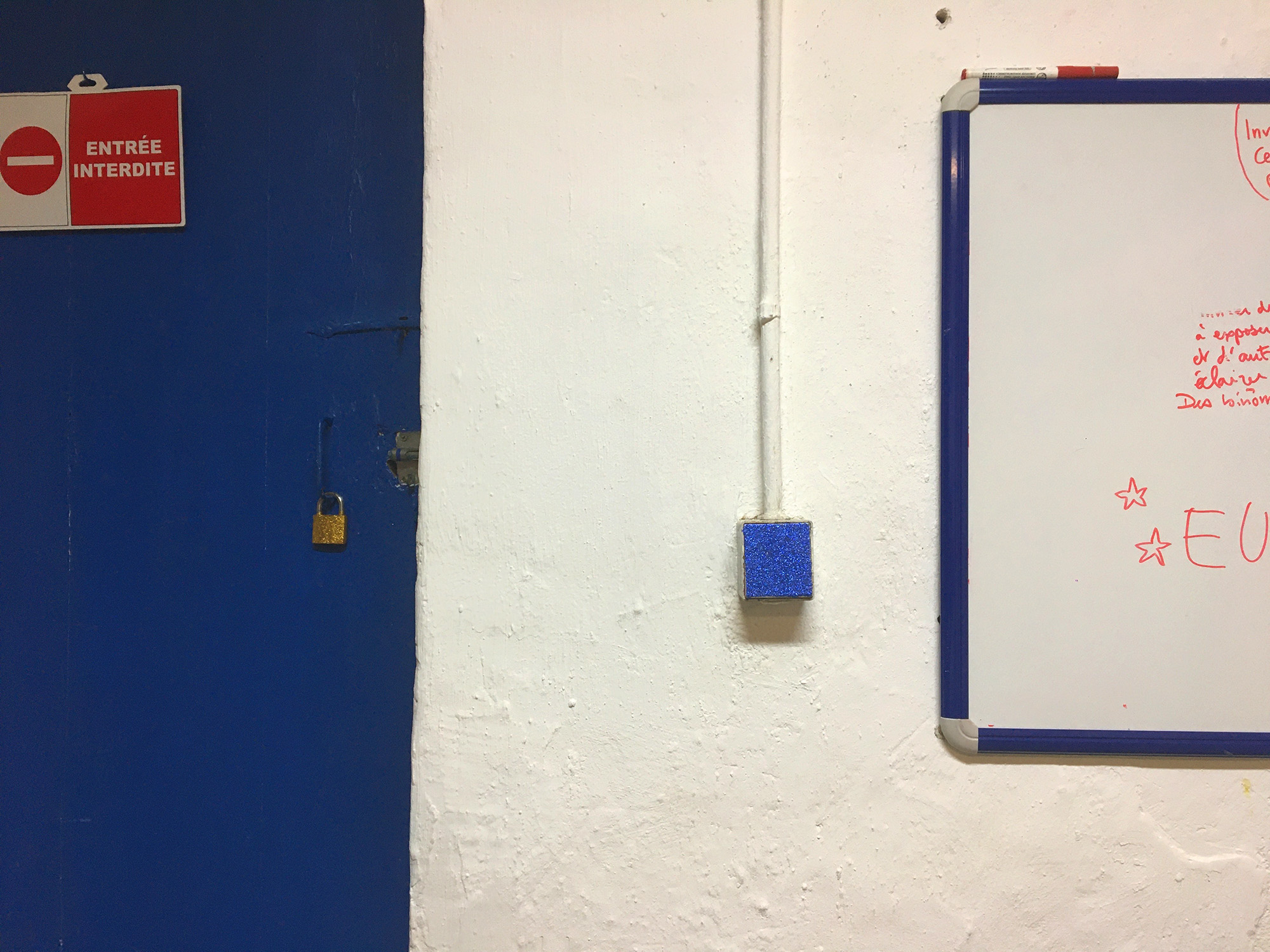 Sur un mur blanc, à côté d'une porte bleu, un interrupteur a été recouvert de paillettes bleues.