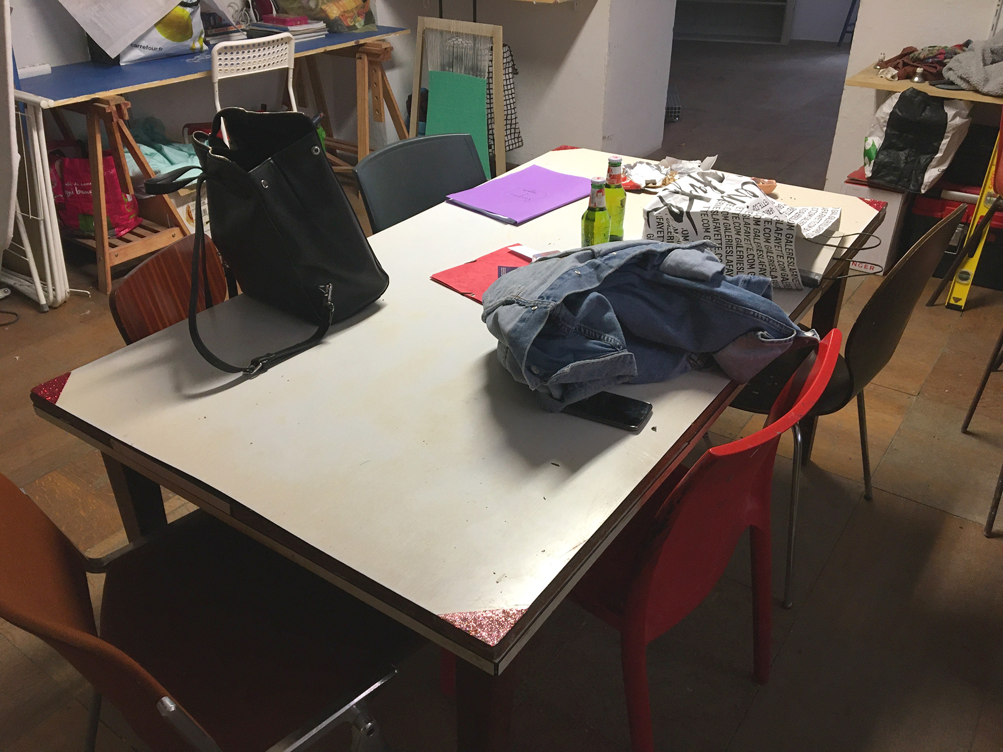 Une table au milieu d'une salle avec des chaises autour et des affaires posés dessus. On voit que les angles de la tables ont des paillettes collées.