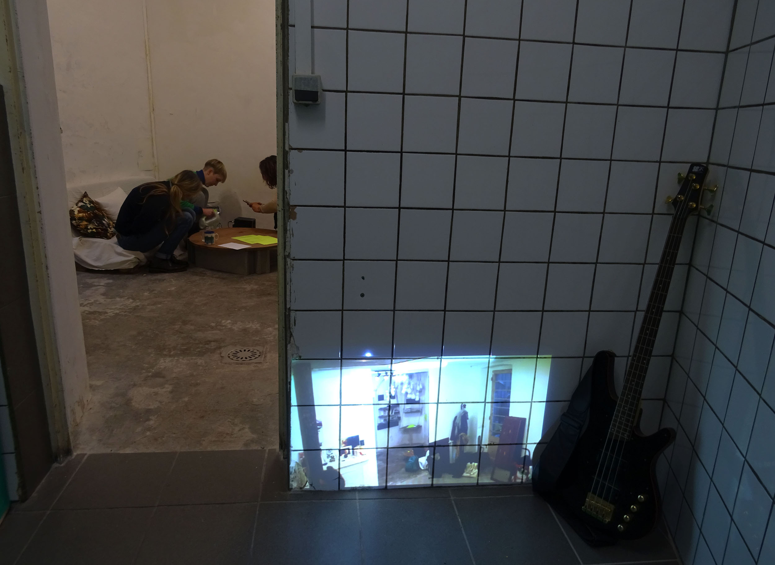 Dans un espace en carelage (mur et sol) on voit une image projeter sur le mur depuis un vidéoprojecteur posé à même le sol. Ce sont les images de l'exposition que l'on apperçoit dans la salle d'à côté aoù des gens sont assis.