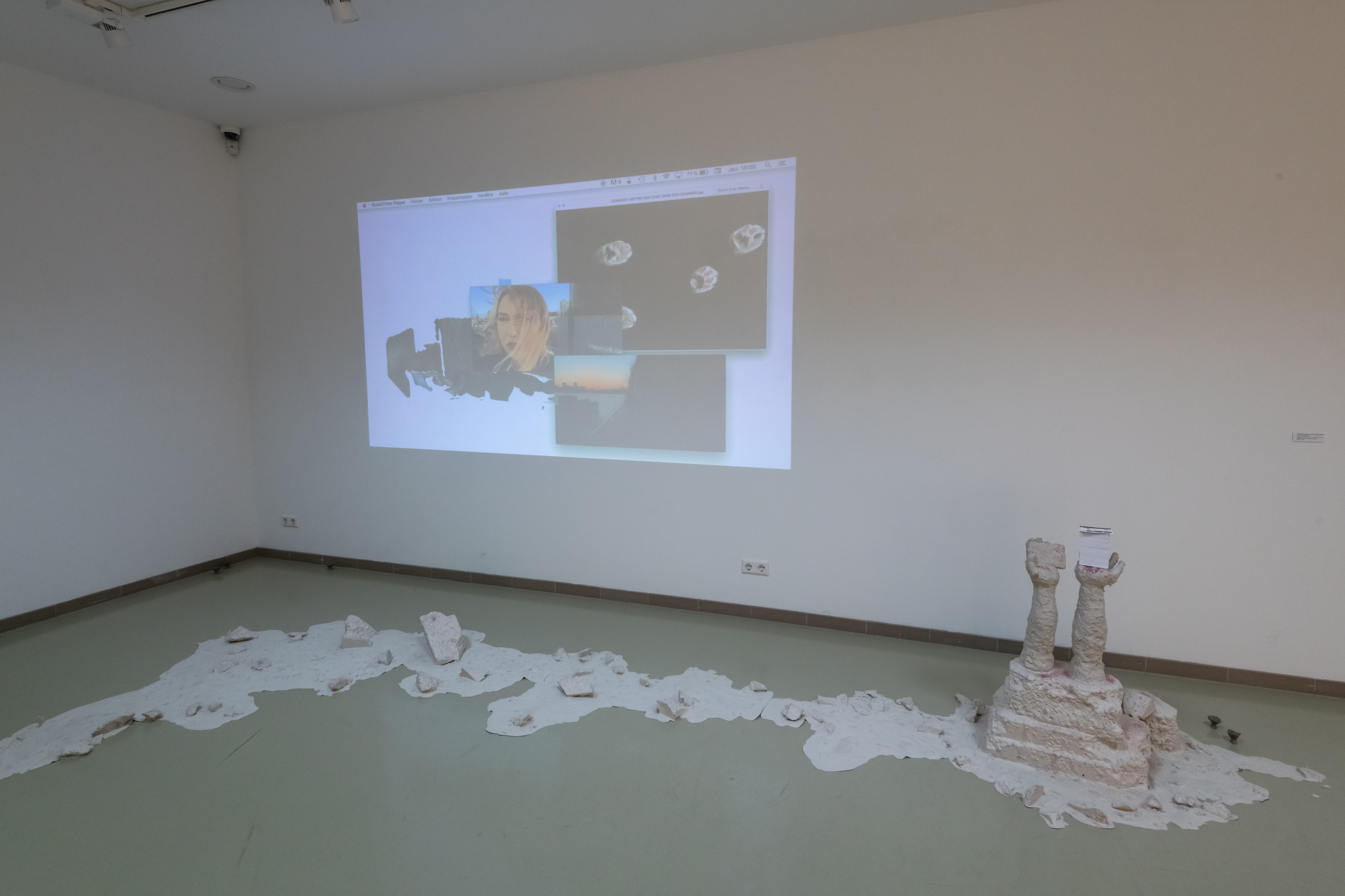 vue de l'installation qui en plus de la projection au mur, dispose d'une sculpture qui ressemble à une flaque de mortier répendu dans toute la salle. Des blocs sont pris dans la flaque et deux mains en sortent. Ces eux mains tiennent les cartes de visite qui reprennent les codes d'une page d'accueil Youtube. 